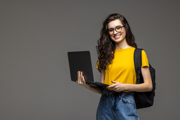 College student girl on gray background, smiling at camera, pressing laptop to chest, wearing backpack, ready to go to studies, start new project and suggest new ideas.