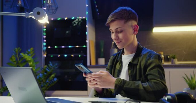 Teenager boy sitting at table holding smartphone enjoying using mobile apps, playing games texting SMS message learning online. Male working remote with technology.