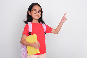 Asian kid wearing school bag and bring a book pointing to the left side