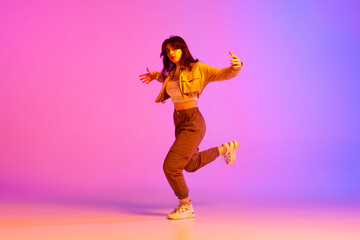 Fototapeta na wymiar Dynamic image of young female dancer in sport style clothes dancing against gradient pink purple background in neon light. Concept of contemporary dance, youth, hobby, action and motion