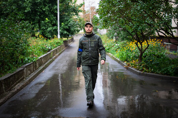 Serious military man in olive uniform and cap walking on street on rainy summer day