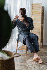 Fototapeta na wymiar African american man with vitiligo in robe drinking coffee on bedroom bench at home.