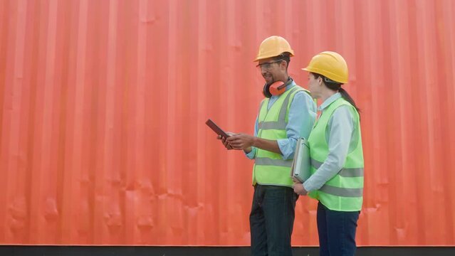 Mixed-raced workers walking near red freight container. Young man using tablet device and headphones. Pretty female holding folder with documents. People with safety equipment actively speaking.