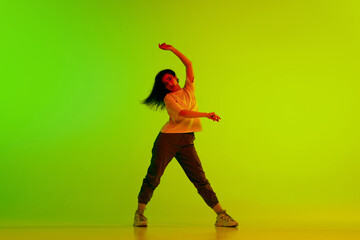 Dynamic image of young girl, female hip-hop dancer in motion against gradient green yellow background in neon light. Concept of contemporary dance, youth, hobby, action and motion