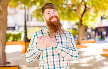 red hair bearded man feeling romantic, happy and in love, smiling cheerfully and holding hands close to heart