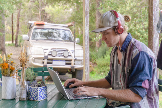 Man working remote on laptop with headphones on in the bush with trees and 4x4 car in background