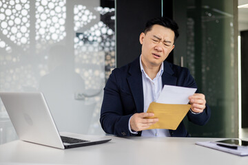 Frustrated and sad depressed businessman received mail envelope notification bad news, asian man at workplace inside office working with laptop, unhappy with achievement results.