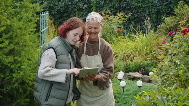Medium shot of modern gen Z girl showing photos on digital tablet to her grandmother while spending time in garden
