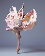Young classical dancer wearing colorful flying dress dancing on fingertips over grey studio background. Graceful ballerina © Lustre Art Group 