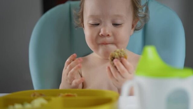 Baby girl sitting in chair splits off piece of meatball and eats it for lunch. Caucasian hungry todler with curly hair eats dinner, bites and chews healthy food with his hands