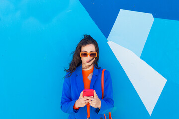 Fashionable young woman using smart phone in front of blue wall