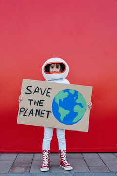 Girl wearing astronaut costume holding Save The Planet placard standing in front of red wall