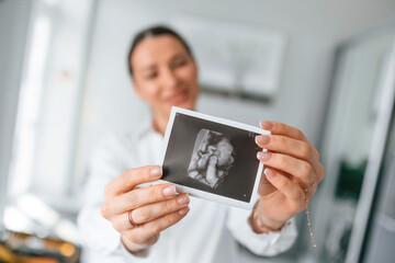 Standing and smiling. Woman in white coat is holding ultrasound picture of baby photo