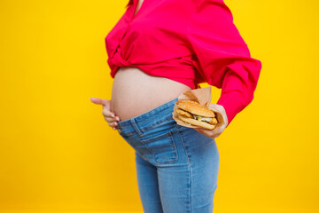 Cheerful pregnant woman in pink shirt holding hamburger over isolated yellow background with surprise and shocked facial expression. Harmful food during pregnancy. A pregnant woman eats fast food