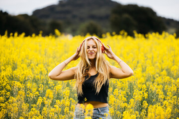 Naklejka na ściany i meble Beautiful blonde young woman listening music with red headphones amidst a field of blooming yellow rapeseed flowers