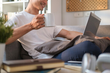 Smiling man in casual clothes hand holding coffee cup ad using laptop on couch.
