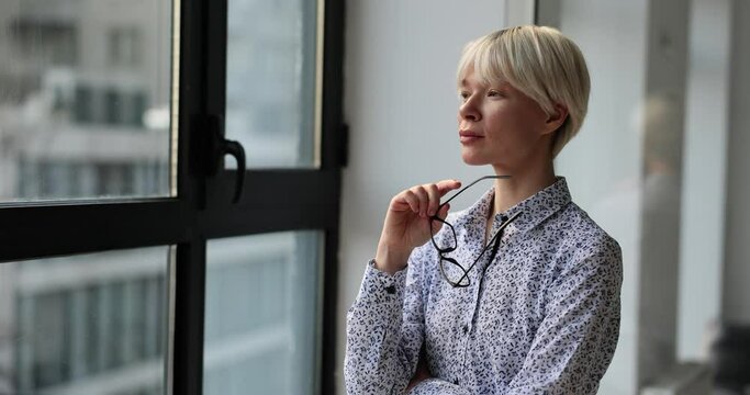Thoughtful blonde woman looks out of window holding stylish glasses in corporate building. Lady boss plans business project in office
