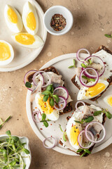 Smorrebrod sandwiches with herring fillet, rye bread, cream cheese, boiled egg slices, onion rings and microgreens on white dish brown background, rustic style. Top view