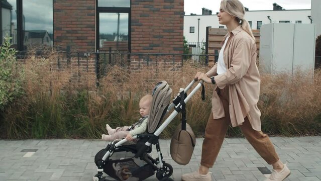 Side view tracking tilt up shot of young mother wearing casual clothes walking along street with her baby in stroller