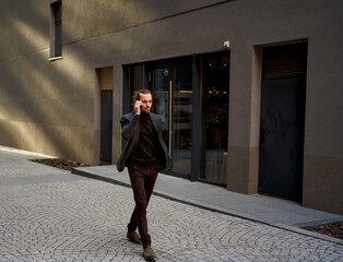 Urrban Image of confident young businessman in black suit walking on the street while busy talking on the phone call. Perfect for commercial projects related to business and electronics.