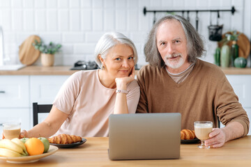 Obraz na płótnie Canvas Happy married caucasian middle aged couple, husband and wife, sit at home in the kitchen, have croissants and coffee for breakfast, use a laptop during breakfast, look at camera, smile