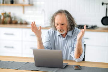 Frustrated caucasian gray haired elderly man, pensioner, financial mentor, sits at a table in the kitchen, looks confusedly at a laptop, is perplexed by the news or the result of work, gesturing hands