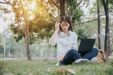 Portrait of young asian woman in white shirt sitting on green grass in park with legs crossed during summer day while using laptop and wireless headphones for video call.