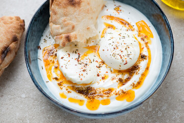 Bowl of turkish poached eggs served in yogurt and topped with melted spicy butter, middle closeup on a beige stone background - obrazy, fototapety, plakaty