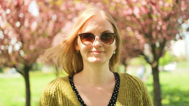 Cute blonde woman is enjoying a spring blossom. Happy smiling girl looking at the camera. A sunny spring in the fresh air. Spring blooms of a sakura tree in a city park. Super slow motion photography.