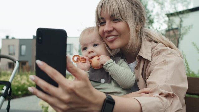 Selective focus shot of cheerful young Caucasian woman sitting outdoors with baby on her lap chatting on video call on smartphone
