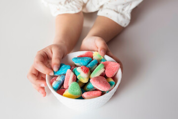 Child hands holds a bowl with colorful sour and sweet candies on a white background.