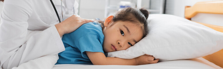 Obraz na płótnie Canvas sick and frustrated asian child looking at camera while lying on hospital bed near doctor, banner.