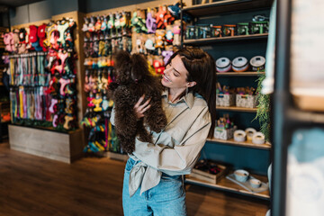 Beautiful young woman enjoying in modern pet shop together with her adorable brown toy poodle.