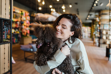 Beautiful and happy young woman sitting in modern pet shop cafe bar and enjoying in fresh coffee together with her adorable brown toy poodle.