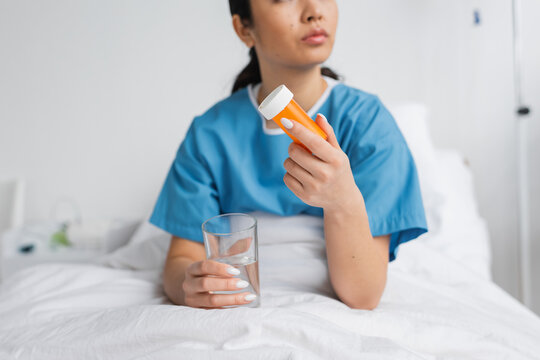 partial view of woman in hospital gown holding water and pills container on bed in clinic.