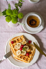 Belgian waffles with bananas and cherries on a white plate, cup of tea and flowers. Sweet breakfast