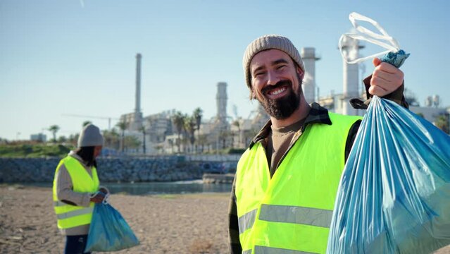 Happy volunteer recycling showing his plastic trash bag looking at camera. Smiling ecology activist cleaning the beach and the sea of pollution and waste, collecting the garbage on a bin. Slow motion - Powered by Adobe