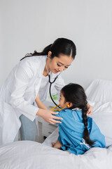 smiling asian pediatrician with stethoscope examining child in hospital gown sitting on bed in clinic.