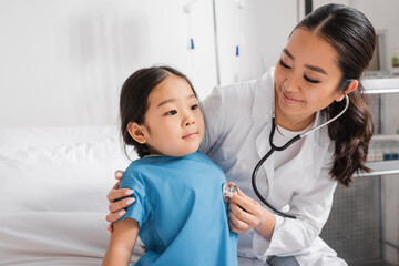 joyful doctor examining little asian girl with stethoscope in pediatric clinic.