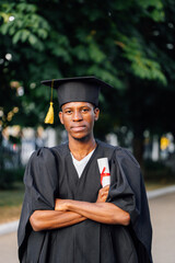 Smiling afro american graduate from university stand outdoors in black mantle and hat with crossed arms and holding higher education diploma, selective focus. Graduation, ceremonial celebration.