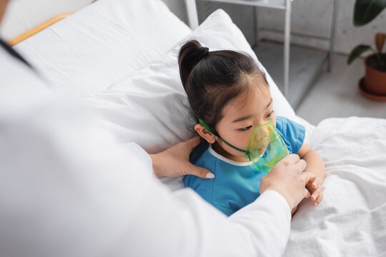blurred doctor adjusting oxygen mask on sick asian child on hospital bed.