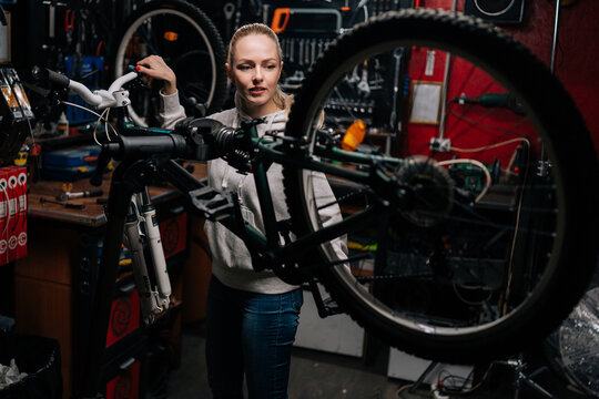 Professional blonde cycling mechanic female watching rear shifter of mountain bike, changing speeds using handlebar shift lever, working in bike repair workshop with dark interior, bike maintenance.