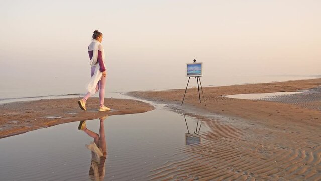 Elegant woman with hair bun walks along sand spit to easel