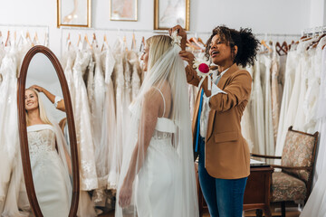 A Multiracial tailor is putting a veil on a happy Caucasian bride in a bridal salon.