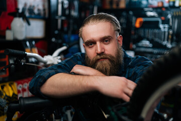 Closeup face of bearded cycling repairman standing leaning on bicycle in repair bike workshop with dark interior, serious looking at camera. Concept of professional repair and maintenance of bicycle.