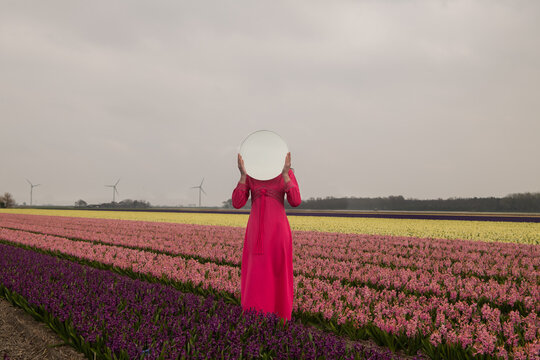 woman in pink dress standing in a field of tulips holding round mirror