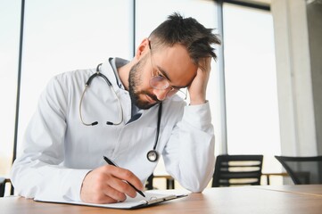 Handsome doctor man wearing medical uniform sitting on his workplace tired holding his head feeling...