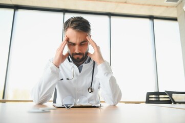 Handsome doctor man wearing medical uniform sitting on his workplace tired holding his head feeling fatigue and headache. Stress and frustration concept