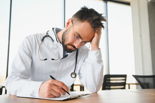 Handsome doctor man wearing medical uniform sitting on his workplace tired holding his head feeling fatigue and headache. Stress and frustration concept