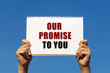 Our promise to you text on notebook paper held by 2 hands with isolated blue sky background. This message can be used as business concept about promise.